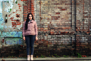 Young cute pregnant girl in pink warm down jacket looks away.  Late autumn, the cold is coming. Old brick wall on the background. toned photo.