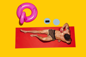 Top view of young caucasian male model's resting on beach resort on red mat and yellow background. Man's lying under sunlight and drinking cocktail. Concept of summertime, party, chill, vacation.