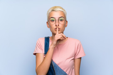 Teenager girl with overalls on blue wall doing silence gesture
