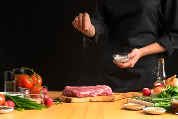 Chef salts steak grill pan. Preparing fresh beef or pork. Horizontal photo with a dark black background.