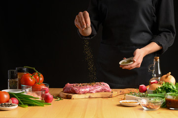 Pork steak and green herbs from the chef in the background. Horizontal photo. With vegetables, tomatoes, hot peppers, onions and a chapel. With black background