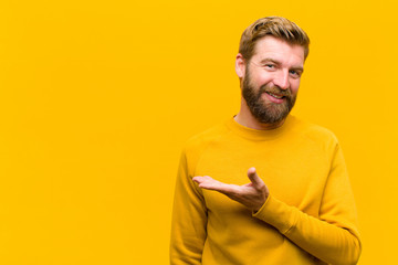 young blonde man smiling cheerfully, feeling happy and showing a concept in copy space with palm of hand against orange wall - obrazy, fototapety, plakaty
