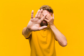 young blonde man covering face with hand and putting other hand up front to stop camera, refusing...