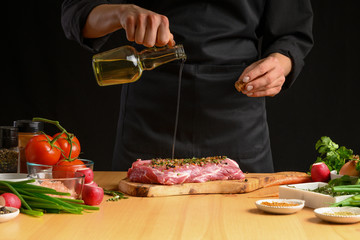 Chef cooks pork steak. pouring oil on the background with vegetables. Recipe book, cooking, restaurant business