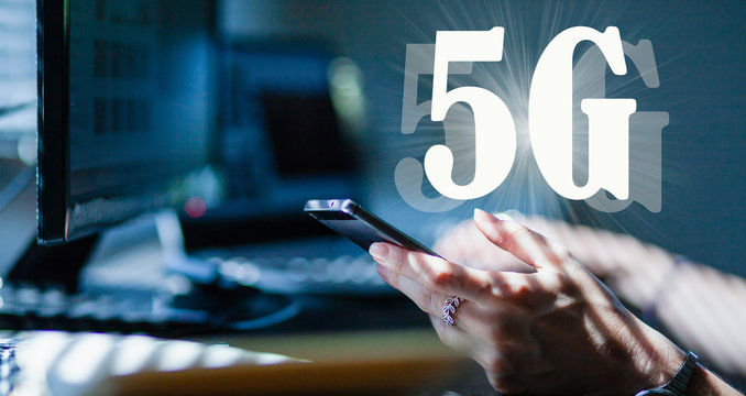 woman working in an office or home office using 5G technology - hand holding smartphone, laptop and monitor in the background