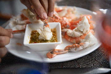 Close up of hand holding steam blue crab and prawn with chili sauce on white plate.