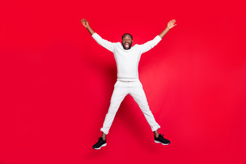 Full size photo of excited dark skin handsome guy jumping high rejoicing having best x-mas party time wear white sweater trousers isolated red background