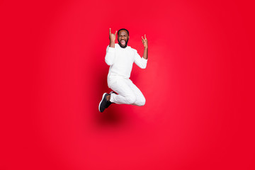 Full size photo of excited dark skin handsome guy jumping high rejoicing at rock concert showing horns hands wear white sweater trousers isolated red background
