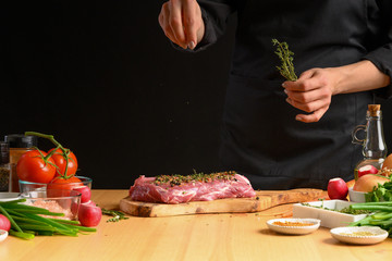 Pork steak and green herbs, rosemary and thyme from the chef in the background. Horizontal photo. With vegetables, tomatoes, hot peppers, onions and a chapel. With black background