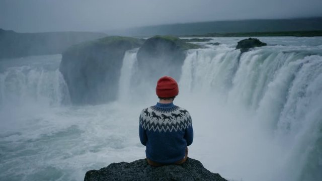 Authentic original shot of romantic and inspiring young man in traditional wool sweater and beanie hat, sit on edge of rock cliff overlook waterfall. Thoughtful and majestic cinematic experience