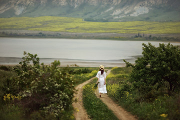 A girl in a white dress, in an elegant hat with a bouquet of yellow field flowers walks down a rural road in the middle of green meadows. Amid mountains and lakes. Provence, passage.