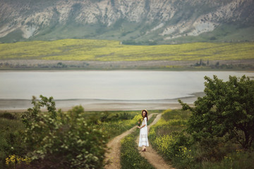 Fototapeta premium A girl in a white dress, in an elegant hat with a bouquet of yellow field flowers walks down a rural road in the middle of green meadows. Amid mountains and lakes. Provence, passage.