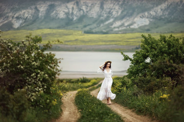 A girl in a white dress, in an elegant hat with a bouquet of yellow field flowers walks down a rural road in the middle of green meadows and laughs. Against the background of mountains and lakes