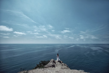 A beautiful girl, a bride, in a white dress, stands the back of on a rock against the background of the sky and ocean. Fashion, style, wedding. Space.