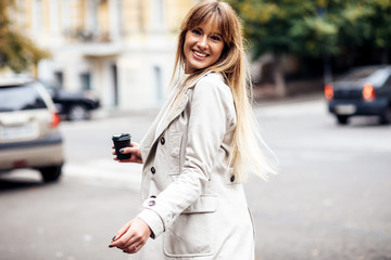 Fototapeta premium The concept of street fashion. portrait of Young girl dressed in beige trench coat. Posing against the window of the boutique girl smiling and drinking coffee