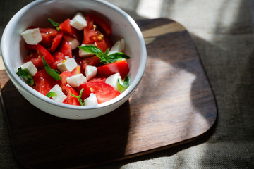 Sliced tomatoes in white bowl with basil leaves and slices of feta cheese. On a wooden board On a linen tablecloth. Horizontal orientation.