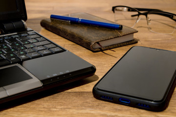 Remote workplace. Remote working environment. View of a natural wood table with several items, a black laptop, a diary, a pen, glasses and a mobile phone.