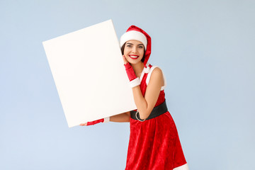 Beautiful young woman in Santa Claus costume and with blank poster on light background