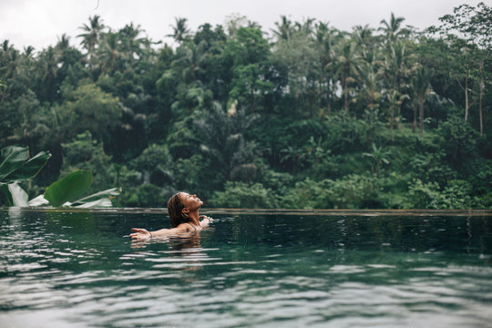 Woman enjoying tropical rain while swimming in infinity pool on Bali
