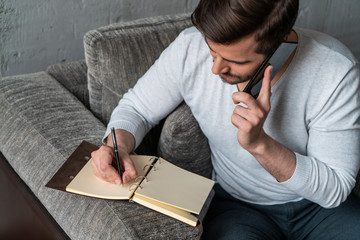 Top view of handsome bearded businessman in casual clothes talking on smartphone and writing in...