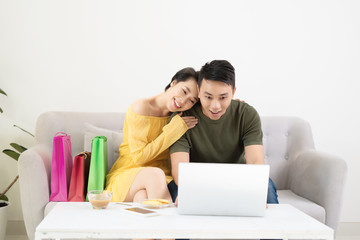 Young Couple Shopping Online On Laptop With Multi Colored Shopping Bags On Sofa