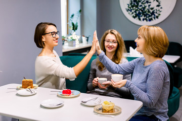 Three girl friends drinking coffee and eating desserts in the cafe. Two women giving five each other, third woman holds cup of coffee and smiles.