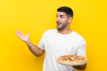 Young handsome man holding a pizza over isolated yellow wall with surprise facial expression