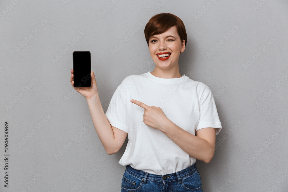 Wall mural Image of lovely young woman smiling and pointing finger at cellphone isolated over gray background