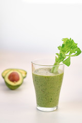 Green smoothie with a stalk of celery in a glass cup. On the table is an avocado.