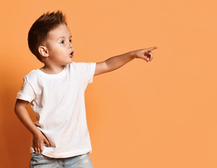 Excited kid boy in white t-shirt with his hand in his pocket is pointing at something on free copy space on yellow