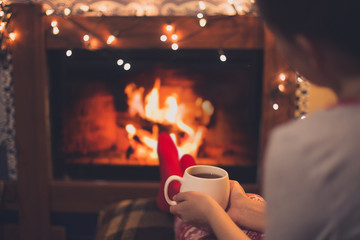 Close up cup of hot tea in woman's hands sitting near fireplace with festive Christmas lights in...