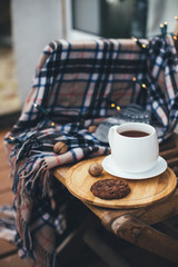 Still life with hot tea and chocolate cookies on wooden tray with warm blanket and festive lights on background. Cozy wooden terrace of a countryhouse. Slow life concept.