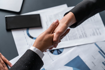 Top view of two businesswomen shaking hands at blurred office table with graphs. Concept of partnership and collaboration