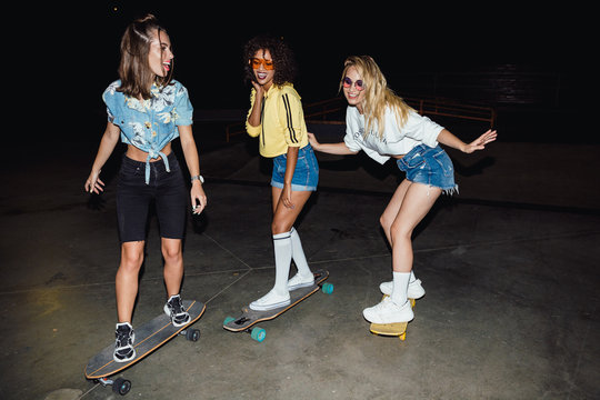 Image of stylish multinational girls riding skateboards at night outdoors
