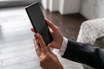 Close up of African American woman hands holding smartphone while she is sitting in armchair in blurred room. Concept of technology and internet