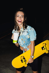 Image of brunette girl drinking soda and holding skateboard at night © Drobot Dean