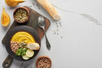 top view of autumnal mashed pumpkin soup on wooden cutting board with spoon and ingredients on marble surface