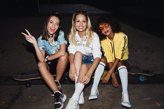 Image of attractive multinational girls sitting on skateboards at night