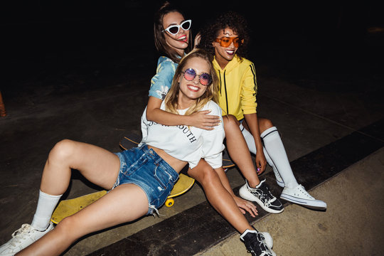 Image of trendy multinational girls sitting on skateboards at night party
