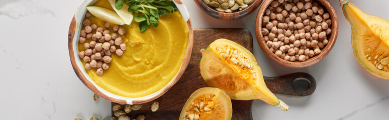 top view of autumnal mashed pumpkin soup in bowl on wooden cutting board near pumpkin, chickpea on marble surface, panoramic shot