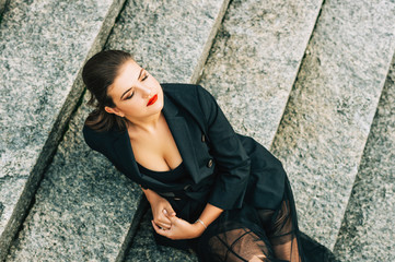 Fashion portrait of beautifl woman wearing black jacket and skirt, dark makeup with red lips, sitting on staire, top view