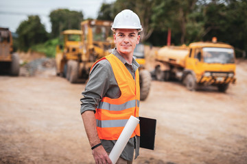 Caucasian handsome engineer is stand and smile holding a laptop with roll of paper on road...