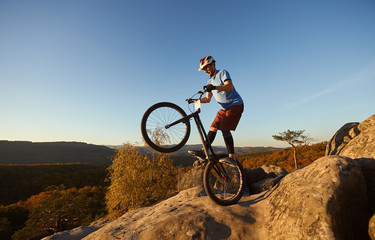 Fototapeta na wymiar Fearless cyclist balancing on back wheel on trial bike. Sportsman rider making acrobatic stunt on the edge of big boulder on the top of mountain at sunset. Concept of extreme sport active lifestyle