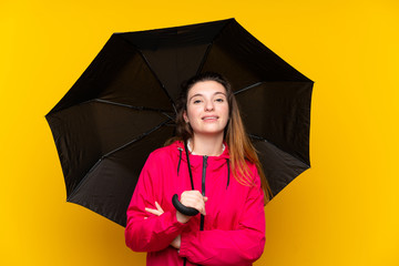 Young brunette girl holding an umbrella over isolated yellow background smiling a lot