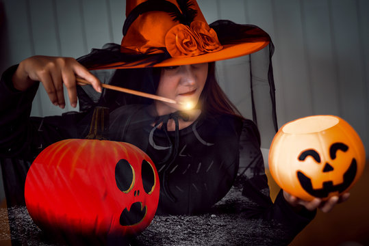 Attractive beautiful Asian woman dressed as a witch using magic wand casting spell to jack pumpkin head, Halloween holidays.