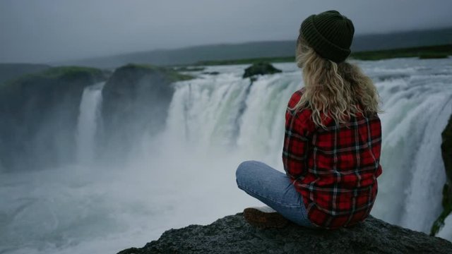 Calm and tranquil young woman enjoys view over epic waterfall during midnight sun in norway or iceland. Scandinavian adventure concept, exploring natural wonders in great outdoors