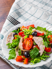 Fresh salad with chicken breast, arugula, black olives,red pepper, lettuce, fresh sald leaves and tomato on a white plate on wooden table background