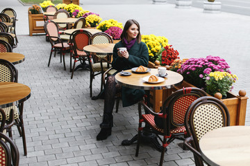Beautiful young girl wearing green coat sitting at a table in cozy street outdoor cafe and drinking...