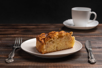 Piece of homemade traditional cornish apple pie on a white plate with a knife and fork on a wooden table. Cup of tea in the background