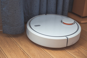 Round white robot vacuum cleaner does its work around the house. Cleaning the house with a robotic vacuum cleaner. Vacuum cleaner cleans difficult places near curtains